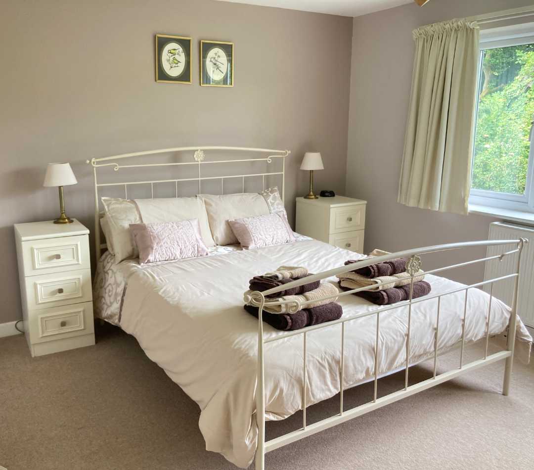 A kingsize metal framed bed with two cream bedside cupboards, a biscuit coloured carpet and some decorative pictures on the walls behind. The bed is made up with cream and gold linen and has two sets of gold and brown fluffy towels. This is the main bed in the family room at The Holt at Church Stretton.