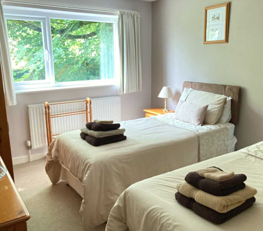 Two twin bed, bedside cupboards and a clothing rack are shown in a light room with a large window. The beds are made up with cream and gold linen and have a set of gold and brown fluffy towels on each. These are the beds in the twin room at The Holt at Church Stretton.