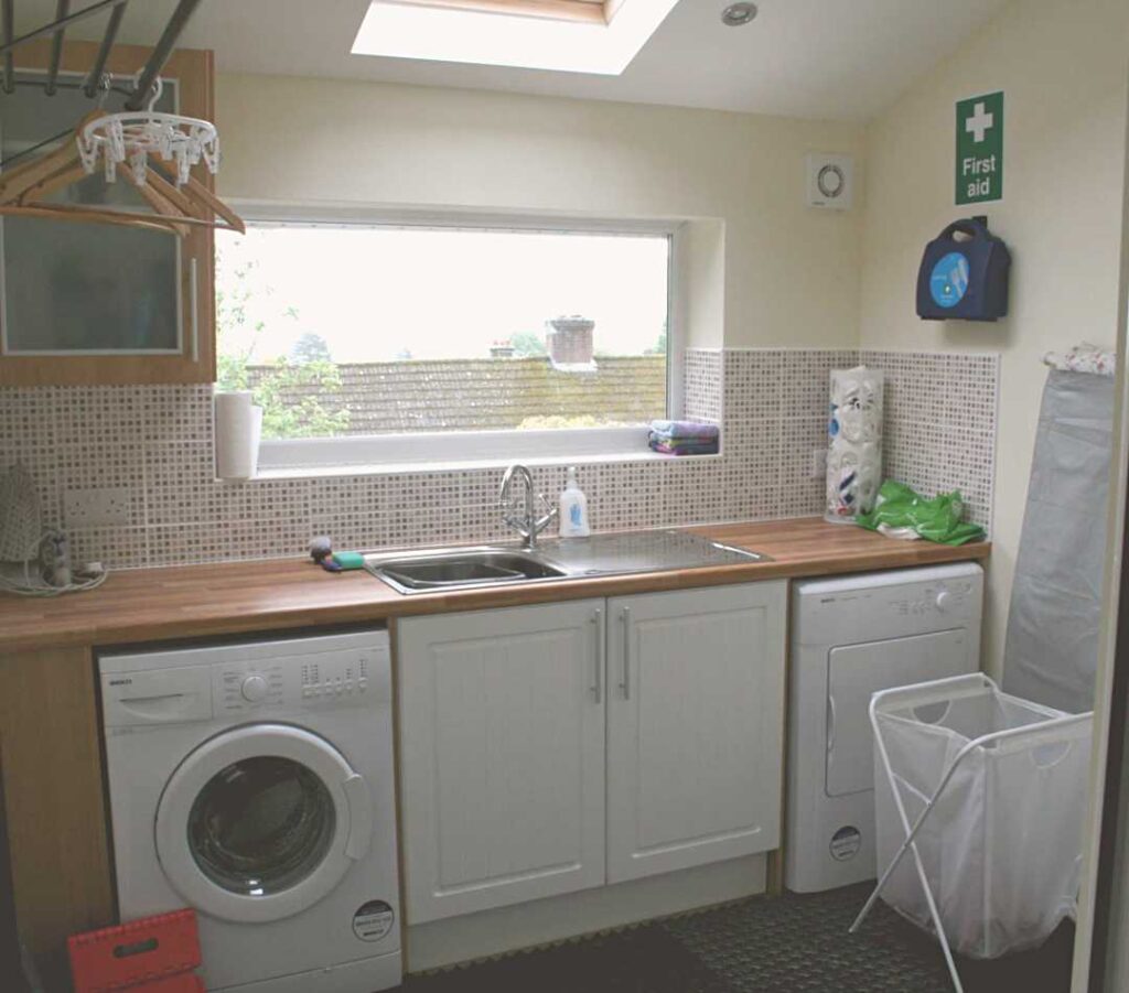 This image shows one side of the utility room at The Holt at Church Stretton. Image shows a washing machine and separate tumble drier, a white under the counter cupboard and a glass wall cupboard, a double sink, an ironing board and washing basket, a drying rack with pegs and hangers and a drip tray underneath, and various cleaning and utility items for use by guests. There is a boot rack slightly out of shot.