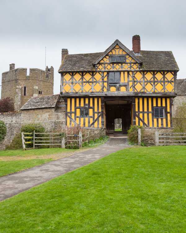 A portait image of the front of the Stokesay Castle manor house, which has tudor frames and yellow plaster, with a castle tower in the background.