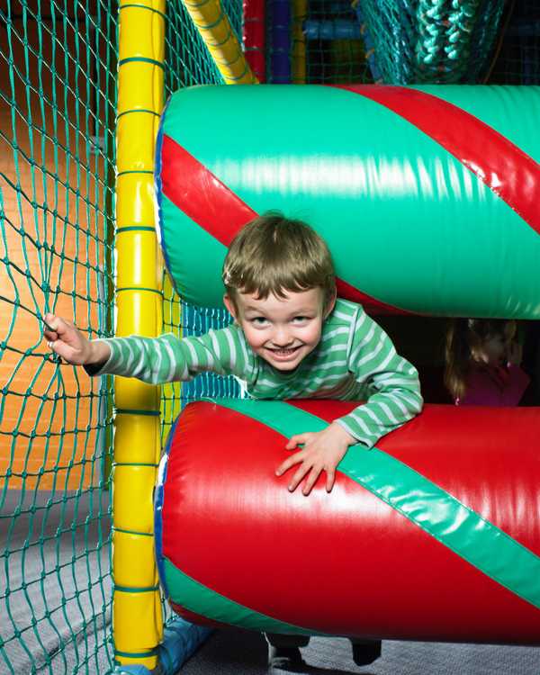 A portrait image representing a soft play centre - image shows a grinning child squeezing between two brightly coloured foam rollers in a netted play setting.