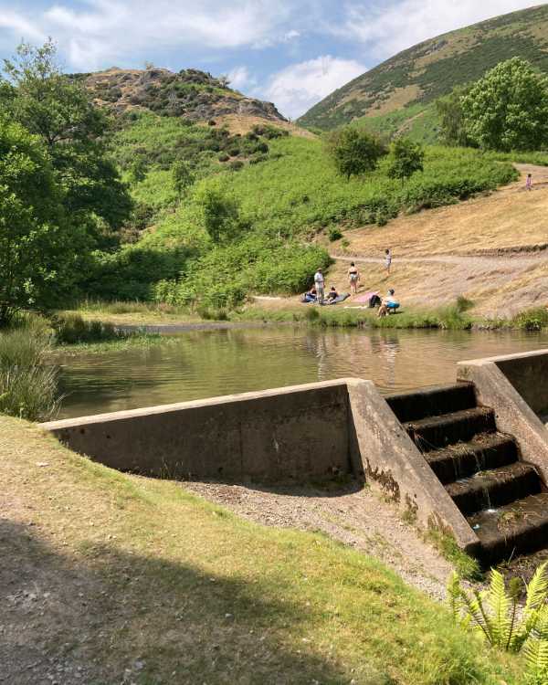 Image of a small reservoir in the Lond Mynd near Cardingmill Valley. Image shows a small square pool with steps funneling the water down into the brook below which leads to the valley. Some families are picnicking in the background and there are hills beyond.