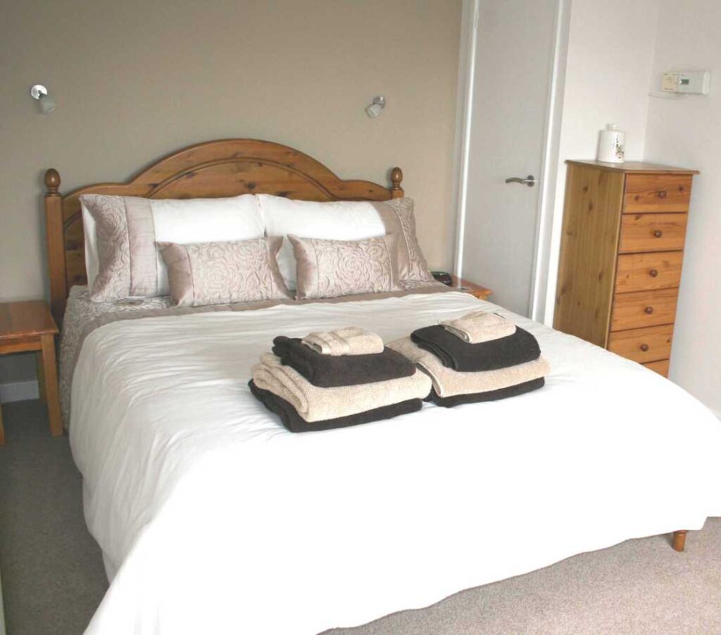A kingsize bed with two bedside cupboards and a chest of drawers, all pine. The bed is made up with cream and gold linen and has two sets of gold and brown fluffy towels. This is the bed in the kingsize room at The Holt at Church Stretton.