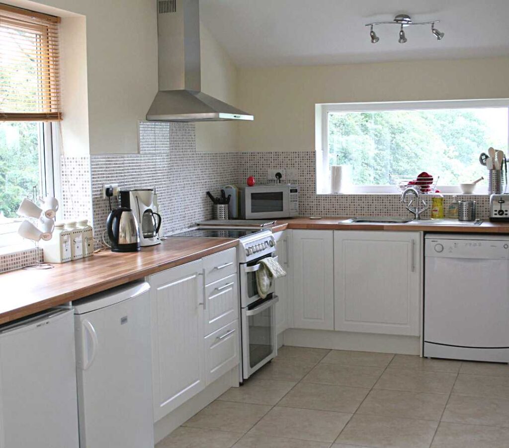 Image of the window sides of the large kitchen diner at The Holt at Church Stretton. The image shows white under counter units and wooden worktops, a fridge, freezer, oven, microwave, sink and dishwasher and some cupboards. The room is light and airy and has two large windows in this image.