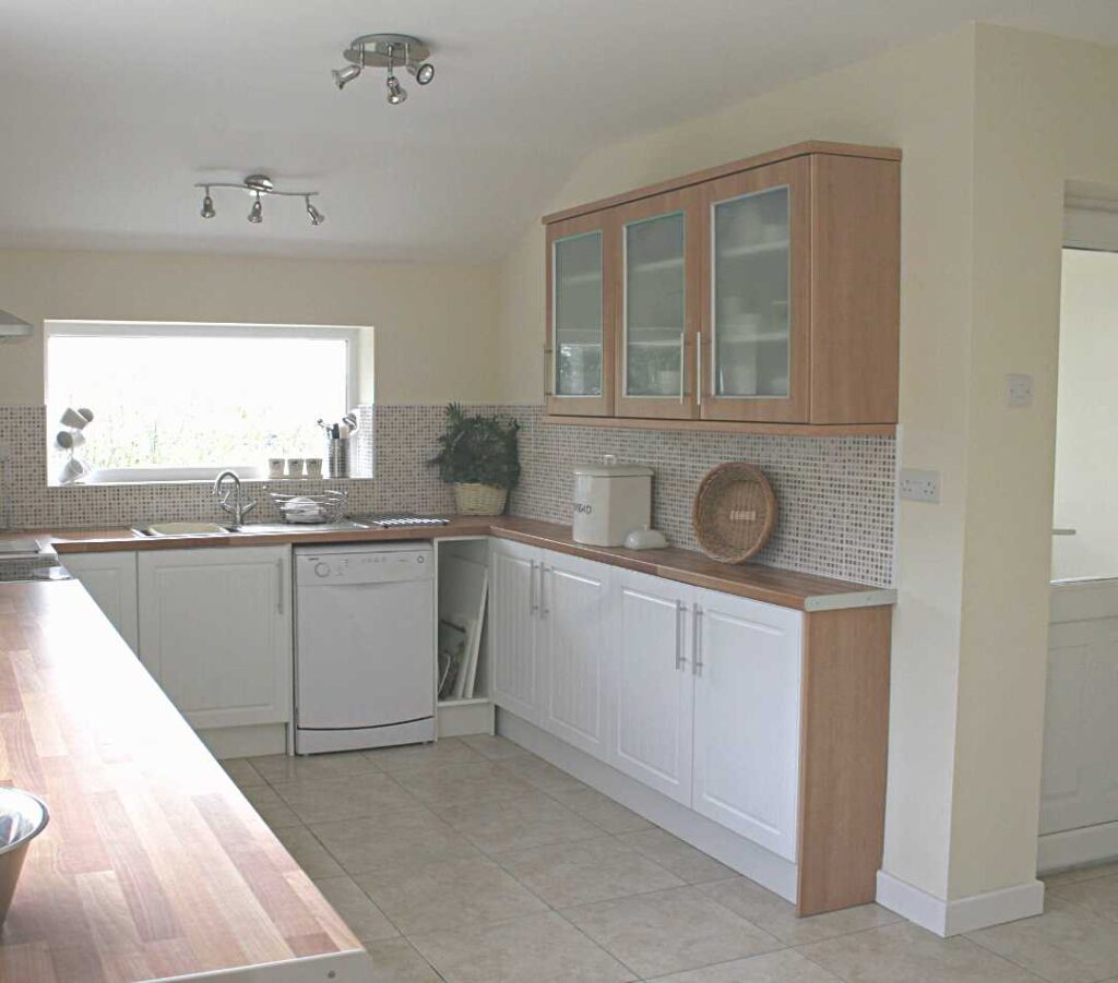 This image shows the utility side of the kitchen at The Holt at Church Stretton. This image includes the dishwasher and sink from the window side photo, and pans round to show more white cupboards under the counter and a wall cupboard full of crockery. You can also see the entrance to a utility room nearby.