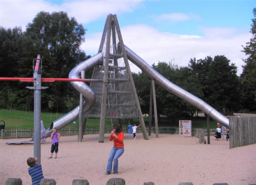 A landscape image representing Telford Town Park, image a huge silver climbing frame with tube slides, and some people using a rotating rope swing in the foreground.