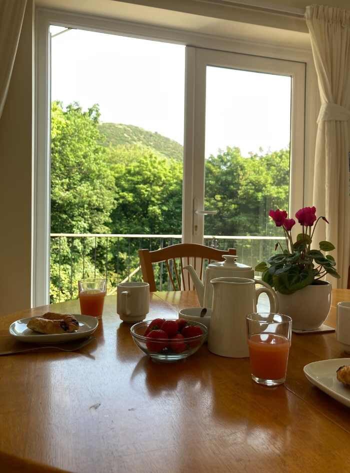 The Kitchen (table close up) at The Holt holiday cottage at Church Stretton, Shropshire This image shows a lifestyle close up of a maple oval table with a breakfast set out on the table, a white coffee set and some pastries and strawberries. Behind are double French windows onto a terrace. You can see that the view on the terrace is of a large hill in The Long Mynd and lots of trees.