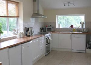 Image of the window sides of the large kitchen diner at The Holt at Church Stretton. The image shows white under counter units and wooden worktops, a fridge, freezer, oven, microwave, sink and dishwasher and some cupboards. The room is light and airy and has two large windows in this image.