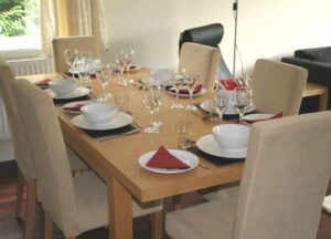 Image shows a beech dining table with six pale cream dining chairs, and place settings for six people, with white china, black mats and red napkins and glassware. A representation of the facilities for a family meal while staying at the Holt.