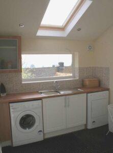 This image shows a portrait view of the middle of the utility room at The Holt at Church Stretton. Image shows a washing machine and separate tumble drier, a white under the counter cupboard and a glass wall cupboard, a double sink, and items for use by guests.