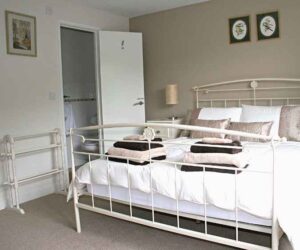 A metal framed kingsized bed and bedside drawers, from an angle which shows a wall with a open door revealing a small ensuite bathroom. The bed is made up with cream and gold linen and has two sets of gold and brown fluffy towels. This is the main bed in the family room at The Holt at Church Stretton.