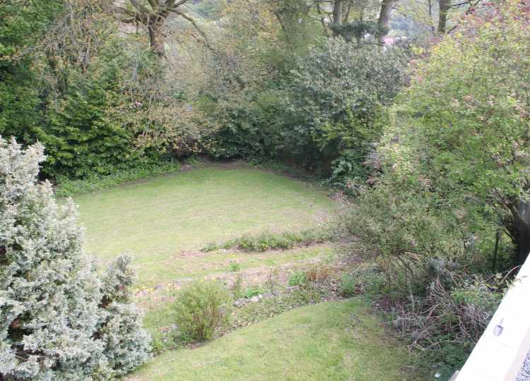 This image shows a garden sloping downwards and to the right from the camera view, with grass terraces and mature trees, taken from The Holt terrace. The garden is very sloping but enclosed and has many mature plants and some flat lawn at the bottom of the slope.