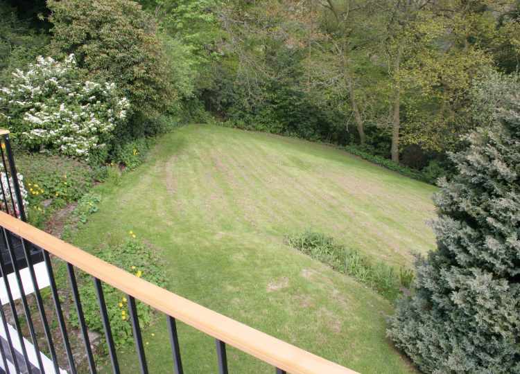 This image shows a garden sloping downwards and to the left from the camera view, with an expanse of sloping lawn and mature trees, taken from The Holt terrace. The garden is very sloping but enclosed and has many mature plants.
