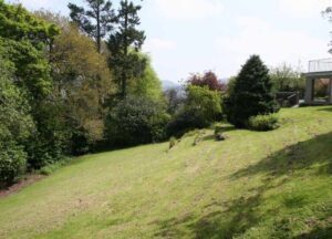 This image shows a sloping garden, taken from halfway up the slope of the garden at The Holt. To the right of shot you can see the terrace of The Holt and the bulk of the image is of lawn and large trees but you can see that there are views to the hills behind the tree line.