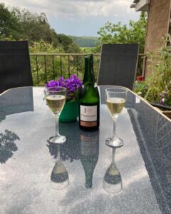 Image shows an open bottle of champagne and two glasses full of bubbles on a glass table with a small purple decorative plant. Behind are chairs and a flower planter. The image is taken on the terrace of The Holt at Church Stretton and the photo shows the terrace fencing and then picturesque views of trees and rolling hills of The Long Mynd in the distance.