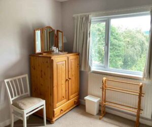 Image of the window end of the Kingsize room at The Holt at Church Stretton. Image shows a pine tallboy with a folding dresser mirror on top, and a glass dish with tissues and hand cream. There is also a chair, a waste bin and a freestanding towel or clothes rack. The image is light because the whole of the back wall is a window with the hint of green trees and hill views showing through.