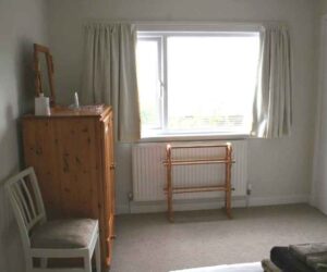 Image of the window end of the Kingsize room at The Holt at Church Stretton. Image is sideways on to a pine tallboy with a folding dresser mirror on top. There is also a chair, a waste bin and a freestanding towel or clothes rack. YOu can just make out the exit door to the right of the window. The image is very bright because the whole of the back wall is a window and it is sunny outside so we can't see the view.