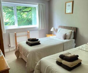 Two twin beds, bedside cupboards and a clothing rack are shown in a light room with a large window. The beds are made up with cream and gold linen and have a set of gold and brown fluffy towels on each. These are the beds in the twin room at The Holt at Church Stretton.