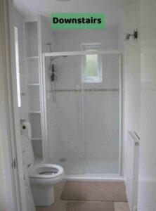 Image shows a bright white and biscuit shower room, with a double sliding shower cubicle, some empty built in shelves, a white toilet and a radiator. There is a label on the image of 