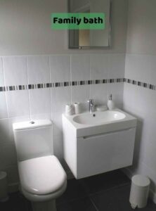 Image shows a white toilet and a white double basin unit with cupboard beneath, and a wall mirror. This is taken with our back to the bath and shower. The floor is dark slate tile. There is a label on the image of 