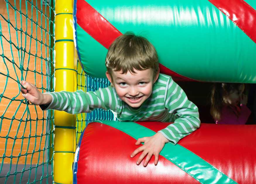 A landscape image representing a soft play centre - image shows a grinning child squeezing between two brightly coloured foam rollers in a netted play setting.