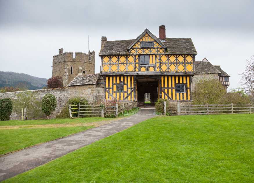 Landscape Image of the front of the Stokesay Castle manor house, which has tudor frames and yellow plaster, with a castle tower in the background.